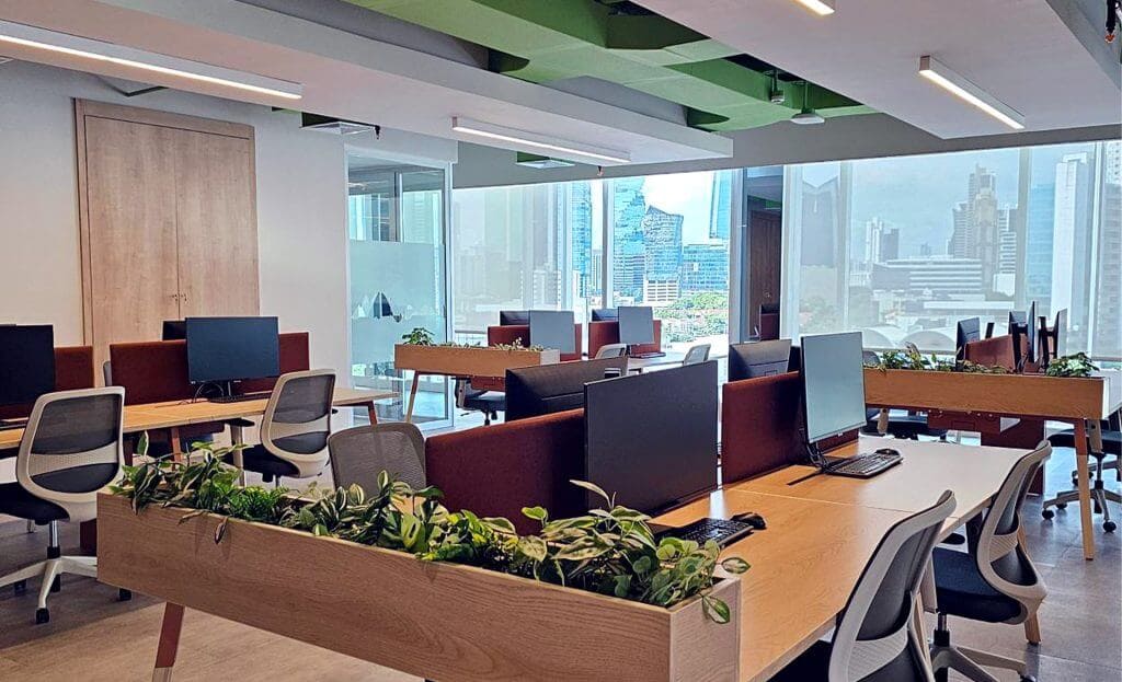 Nosotros 31 Oficina moderna con vista panorámica. Mesas de trabajo compartidas con computadoras y plantas. Ambiente luminoso y acogedor.