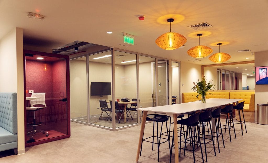 Nosotros 27 Oficina moderna con cabina privada y mesa de trabajo con sillas altas. Iluminación cálida.